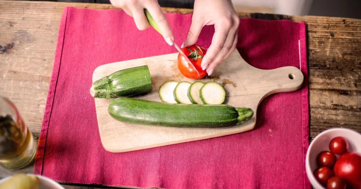 Des mains tranchant une tomate et une courgette sur une planche en bois pour une scène de cuisine vibrante.