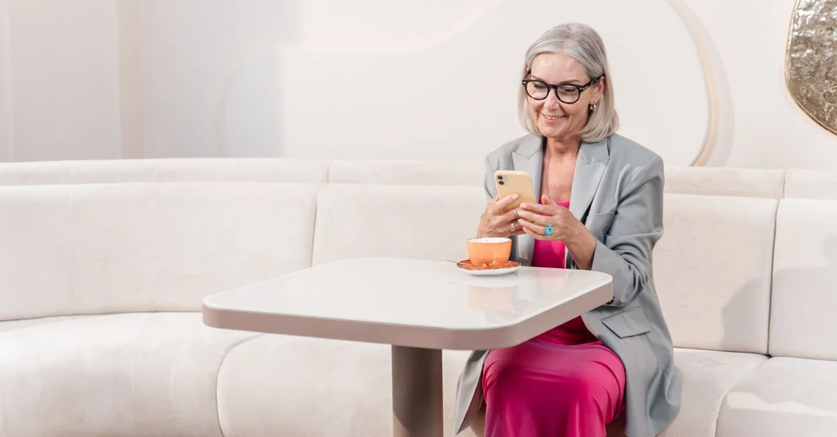 Femme senior utilisant un smartphone à table dans un café, portant des lunettes, souriante.