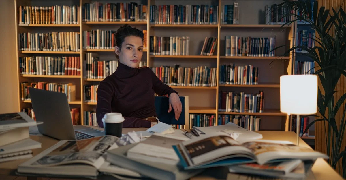Femme travaillant à un bureau avec des livres ouverts et un ordinateur portable dans un cadre de bibliothèque, mettant l'accent sur la recherche et l'étude.