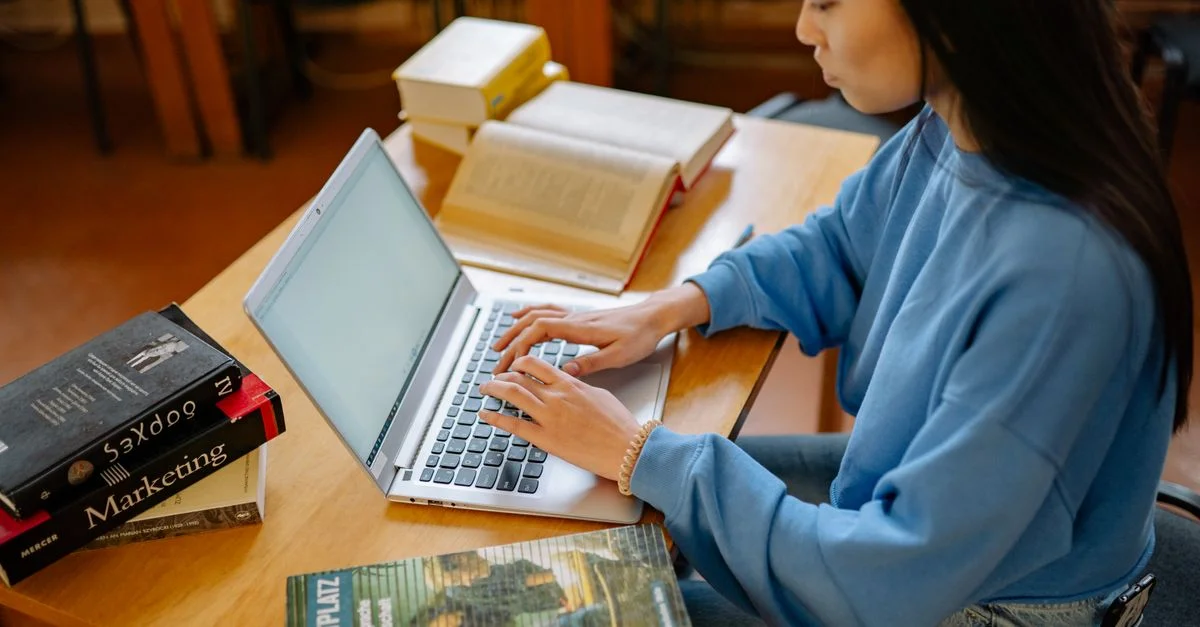 Jeune femme tapant sur un ordinateur portable entourée de manuels dans une bibliothèque.