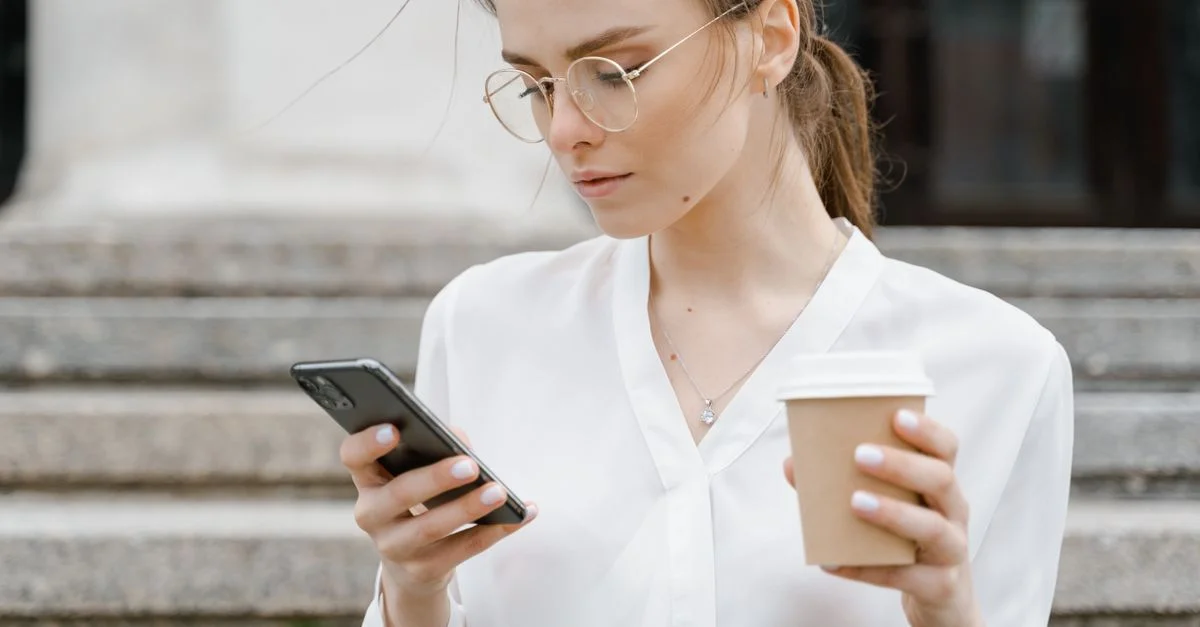 Jeune femme en chemise blanche naviguant sur son smartphone tout en tenant un café à l'extérieur.