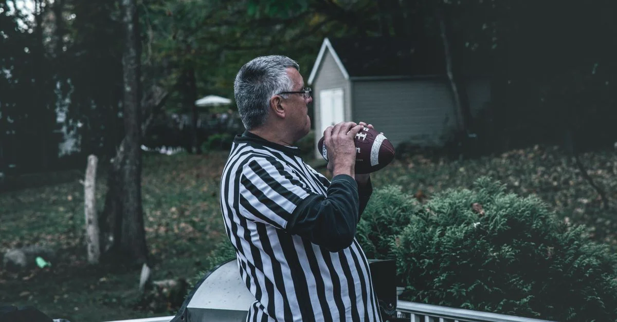 Homme âgé en tenue d'arbitre tenant un football dans un cadre de jardin.
