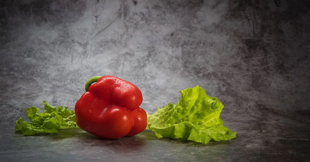 Gros plan sur un poivron rouge frais et des feuilles de laitue croquantes sur un fond gris texturé, soulignant la fraîcheur naturelle.