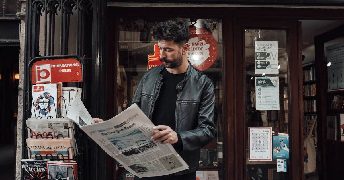 Jeune homme en veste en cuir lisant un journal devant une librairie urbaine.