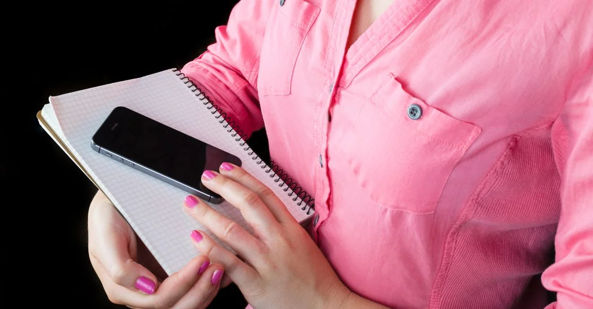 Gros plan d'une femme en chemise rose tenant un smartphone et un carnet, concentrée sur la technologie mobile et la prise de notes.