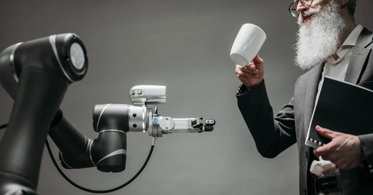 Un ingénieur barbu tient une tasse en céramique tandis qu'un bras robotique assiste, mettant en avant la technologie moderne.