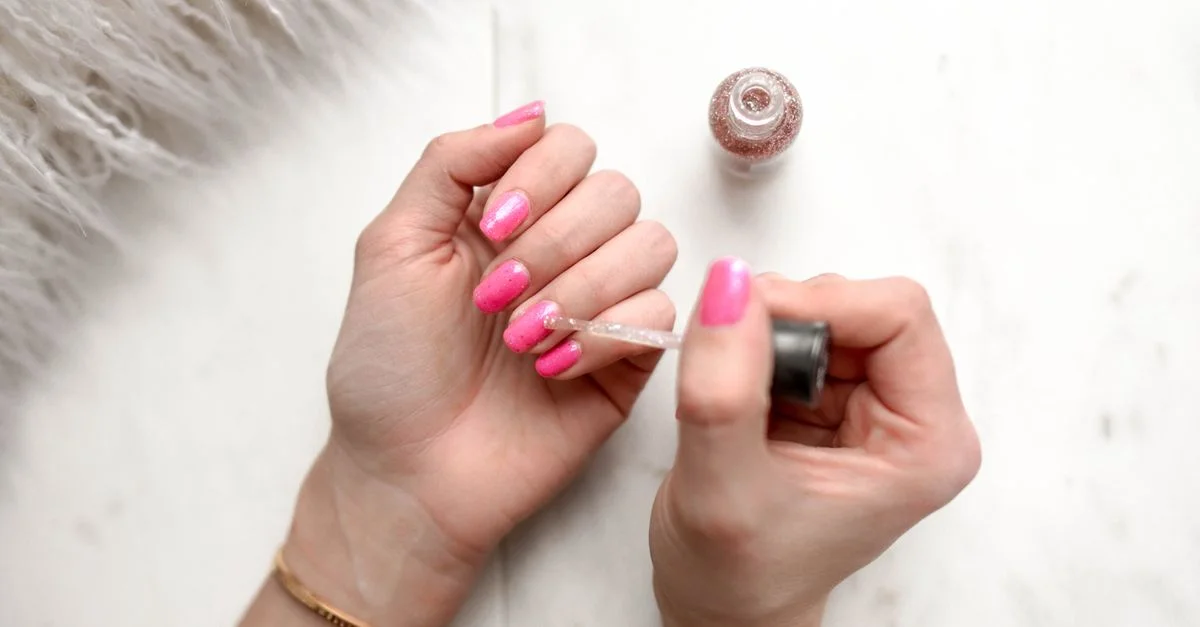 Gros plan sur une femme appliquant du vernis à ongles pailleté sur des ongles roses manucurés.