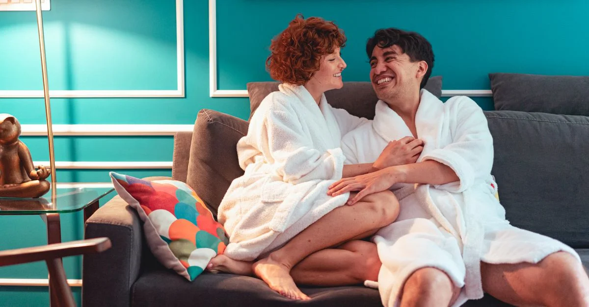 Heureux couple interracial en peignoirs profitant d'un moment cozy sur le canapé à l'intérieur.