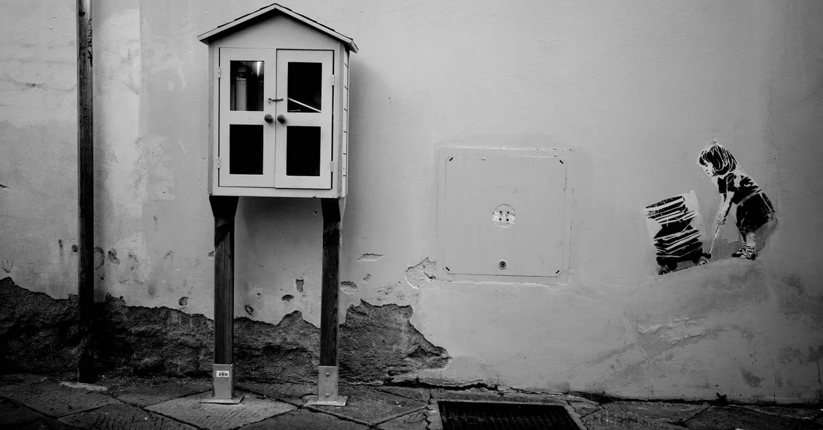 Image monochrome d'art urbain montrant un enfant à côté d'une petite bibliothèque gratuite sur un mur urbain.