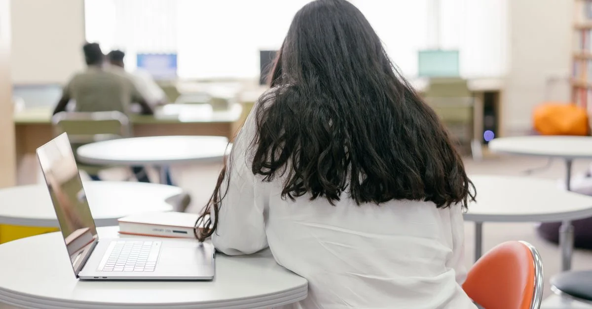 Une étudiante concentrée sur ses études à une table de bibliothèque avec un ordinateur portable et des livres. Atmosphère moderne et éducative.