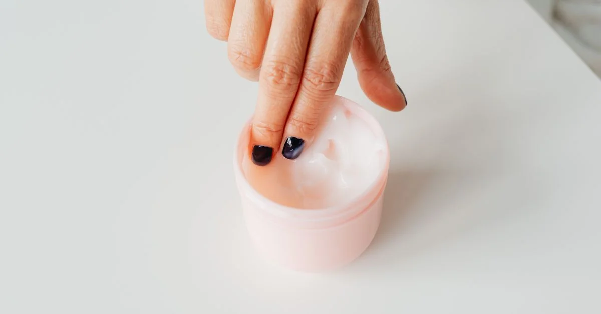 Gros plan sur une main avec des ongles vernis plongeant dans un pot de crème pour le visage, représentant le self-care.