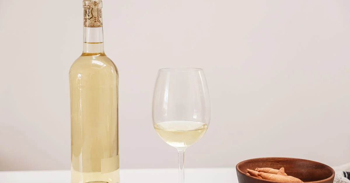 Une bouteille et un verre de vin blanc à côté d'un bol en bois avec des en-cas, posés sur une table minimaliste.