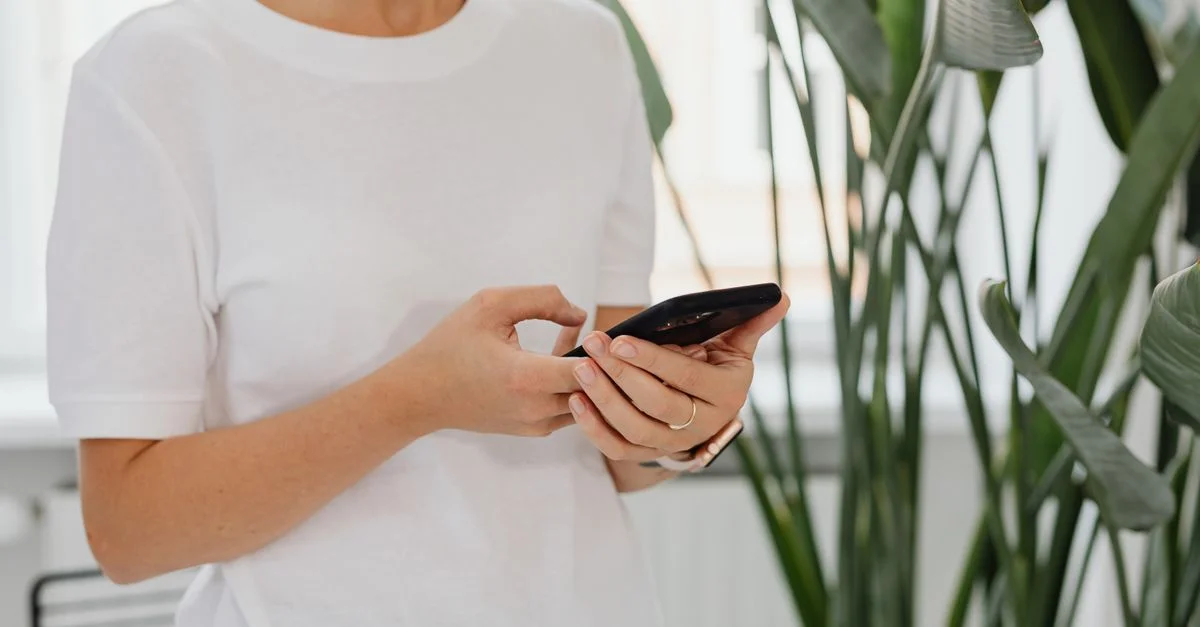 Une femme utilise un smartphone à l'intérieur entourée de plantes vertes luxuriantes.