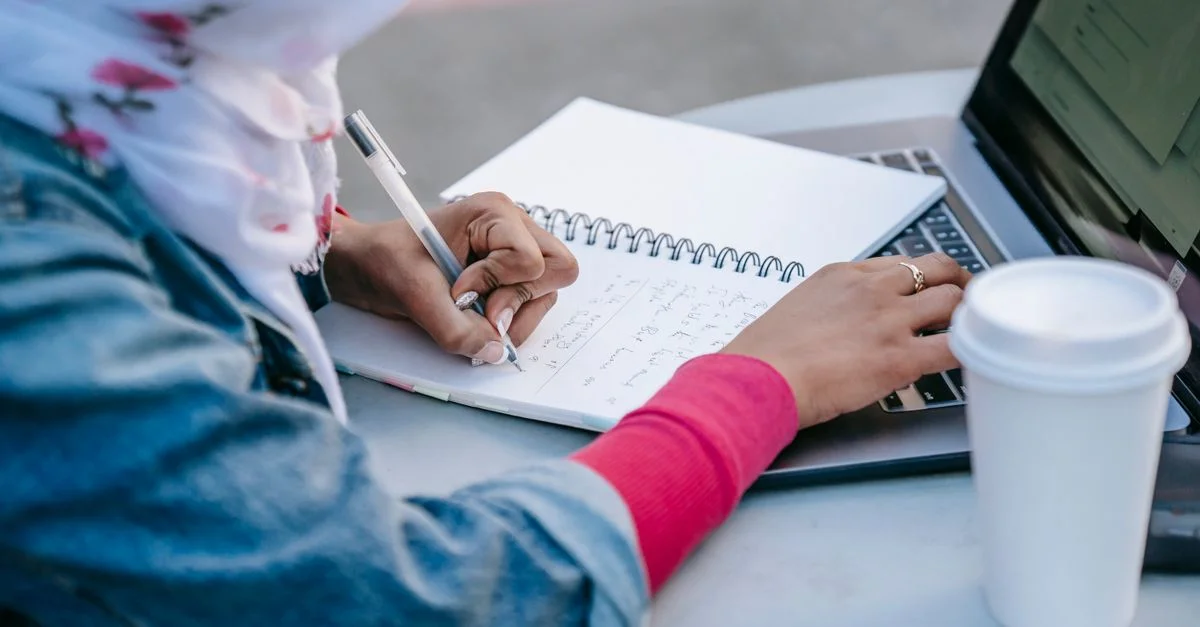 Femme musulmane prenant des notes et utilisant un ordinateur portable en plein air, concentrée sur la recherche.
