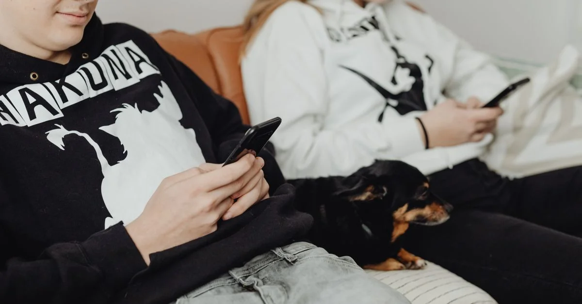 Deux adolescents utilisant des smartphones sur un canapé avec un petit chien. Une scène intérieure détendue illustrant le temps de loisirs.