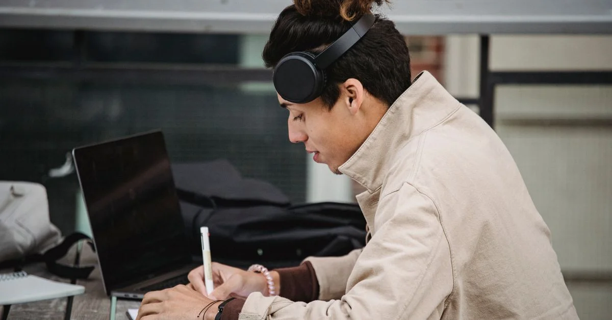 Jeune homme concentré avec des écouteurs étudiant sur un ordinateur portable en plein air, prenant des notes dans un cahier.