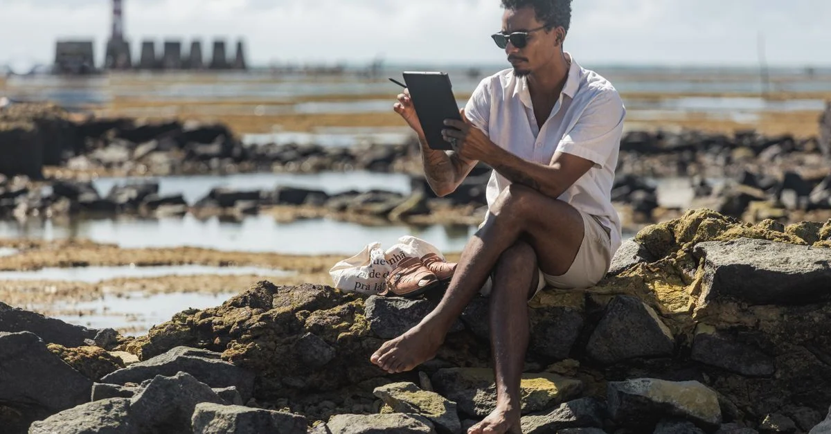 Homme assis sur des rochers près de la plage avec une tablette, profitant d'une journée ensoleillée à Maceió, au Brésil.